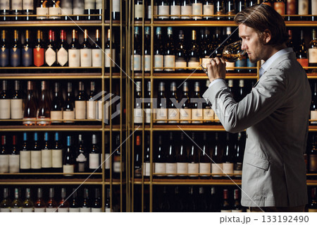 Indoor portrait of experienced male sommelier smells wine in glass standing in cellar over plenty bottle shelf background with a free copy space for your text or logo. Sommelier exam to study wine. 133192490