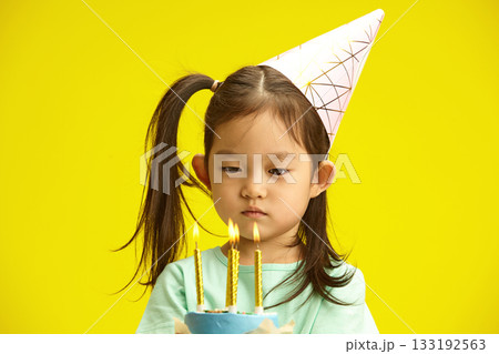 Cute Child Girl Thoughtfully Looking to Cake with Burning Candles, Comes up with Wish that she Wants to Make, Dressed in Festive Outfit, Having Party Hat and Ponytail on Head, Prepare Blow Out Candles Cute Child Girl Thoughtfully Looking to Cake with Burning Candles, Comes up with Wish that she Wants to Make, Dressed in Festive Outfit, Having Party Hat and Ponytail on Head, Prepare Blow Out Candles 133192563