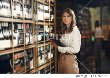 Professional caucasian woman sommelier in wine cellar making notes in tablet and choosing wine from shelve. Association of professional winemaker. 133192566