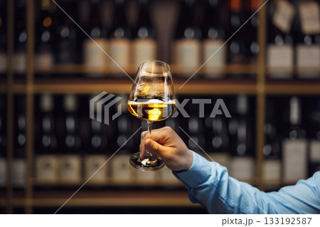Expert Evaluating White Wine Glass Against Blurred Wine Cellar Background 133192587