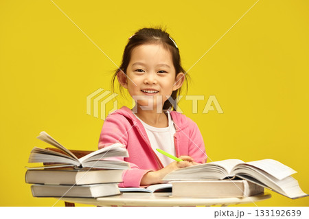 Photo of cheerful little Asian ethnicity first grader schoolgirl enjoying her reading time with books, happy smiling, expression pleasure from studying, sitting at table over vibrant yellow isolated Photo of cheerful little Asian ethnicity first grader schoolgirl enjoying her reading time with books, happy smiling, expression pleasure from studying, sitting at table over vibrant yellow isolated 133192639