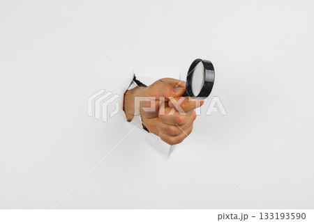 Hand holding magnifying glass emerging from a hole in the wall Hand holding magnifying glass emerging from a hole in the wall 133193590