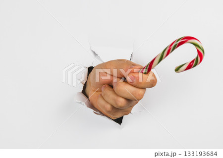 Hand holding a candy cane through a torn hole in a white background Hand holding a candy cane through a torn hole in a white background 133193684