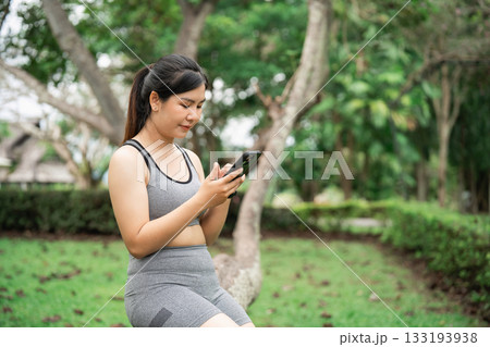 Young woman in sportswear sitting outdoors, using smartphone in nature, practicing wellness, enjoying technology, engaged in physical activity, fitness lifestyle 133193938