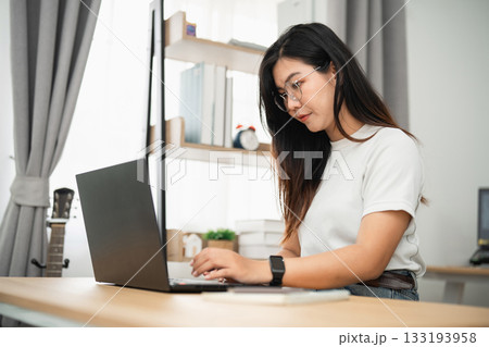 Young woman working on laptop in modern home office, wearing glasses, focused on her task, colorful decor, natural light, creativity and productivity ambiance 133193958