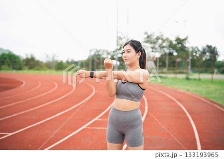 Young woman stretches her arms while preparing for a run on a track, promoting a healthy lifestyle and fitness routine in an outdoor setting 133193965