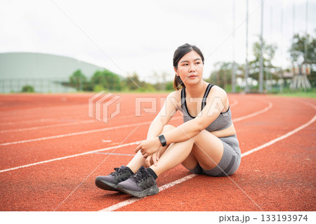 Young athletic woman resting on running track after workout session, wearing sports bra and shorts, focused on fitness and well-being, outdoor training area 133193974