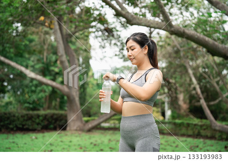 Young woman in activewear drinking water during outdoor workout in lush green park with trees in background, promoting healthy lifestyle and fitness routine 133193983
