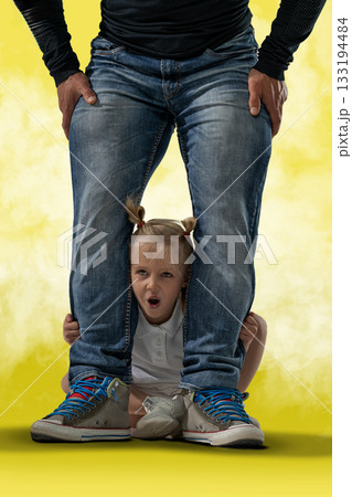 blonde girl with pigtails shows a playful expression while hiding between her father's legs. Her father stands confidently wearing jeans and casual shoes. blonde girl with pigtails shows a playful expression while hiding between her father's legs. Her father stands confidently wearing jeans and casual shoes. 133194484