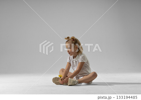 little blonde girl sits on the studio floor looking thoughtfully at a yellow flower in her hand. Soft lighting surrounds her creating a calm and artistic mood. little blonde girl sits on the studio floor looking thoughtfully at a yellow flower in her hand. Soft lighting surrounds her creating a calm and artistic mood. 133194485