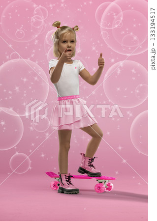 small blonde girl stands confidently on a pink skateboard in a studio setting. She is wearing a white shirt pink skirt and boots. Bubbles surround her in the playful scene. 133194517