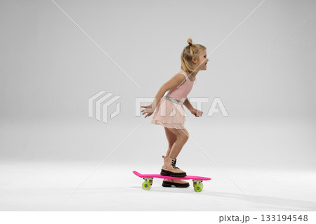 Blonde girl dances on a pink skateboard in a bright studio. She wears a light dress and cheerful expression while balancing effortlessly. Blonde girl dances on a pink skateboard in a bright studio. She wears a light dress and cheerful expression while balancing effortlessly. 133194548