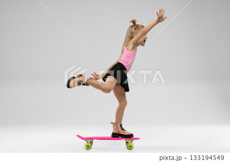 small blonde girl is balancing on a skateboard inside a studio. She is wearing a pink top and a black skirt while having fun and posing confidently with a smile. small blonde girl is balancing on a skateboard inside a studio. She is wearing a pink top and a black skirt while having fun and posing confidently with a smile. 133194549
