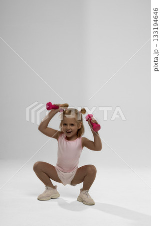 small blonde girl squats in a studio while holding pink dumbbells above her head. She shows excitement and joy while engaging in a fun fitness activity that promotes healthy habits. 133194646