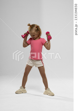 small blonde girl joyfully lifts pink dumbbells. She wears a pink shirt and shorts showing enthusiasm while exercising. 133194650