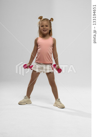 small blonde girl stands confidently with pink dumbbells in each hand. She wears a light pink top and white shorts ready for her workout. small blonde girl stands confidently with pink dumbbells in each hand. She wears a light pink top and white shorts ready for her workout. 133194651