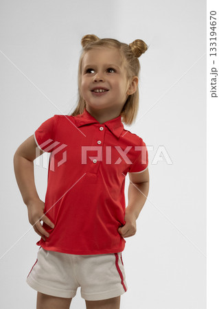 cheerful little girl with blonde hair stands confidently in a studio. She wears a red shirt and white shorts displaying a joyful smile while posing for a picture. 133194670