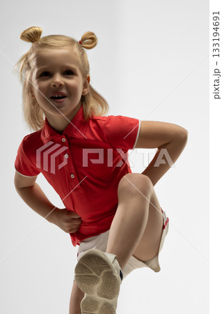 Blonde girl in a red shirt shows a playful pose in a studio. She wears a bright smile while balancing on one leg capturing a moment of joy and creativity. Blonde girl in a red shirt shows a playful pose in a studio. She wears a bright smile while balancing on one leg capturing a moment of joy and creativity. 133194691