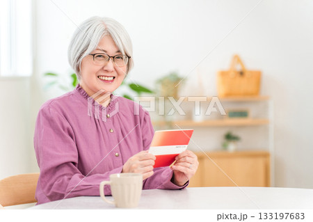 家のリビングで貯金通帳を見る笑顔のシニア女性（老後資金,資産運用,預金,所得,生活費） 133197683