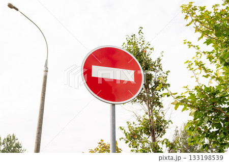 Red no entry sign against bright sky with green trees and streetlamp Red no entry sign against bright sky with green trees and streetlamp 133198539