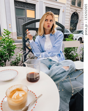 Young blonde woman is sitting at table in cafe, enjoying coffee break and making funny face while holding cup of coffee. Woman sitting at cafe table with coffee and making playful expression 133202005
