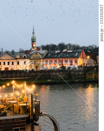Birds flying over Krakow riverside with illuminated restaurant and St. Andrew Church in background at dusk, creating magical atmosphere. Silhouetted birds soaring above Krakow riverbank. Evening City 133202027