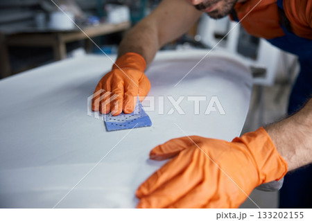 Craftsman sanding surfboard in workshop with hands with orange gloves finishing board Craftsman sanding surfboard in workshop with hands with orange gloves finishing board 133202155