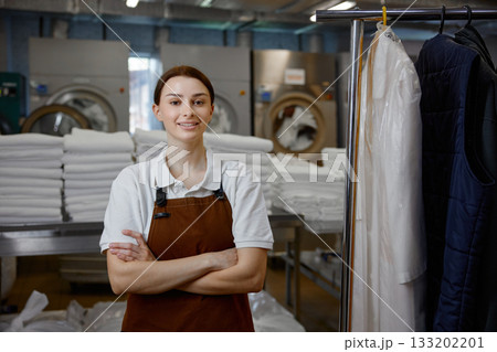 A woman worker in an apron stands with arms crossed in a laundromat A woman worker in an apron stands with arms crossed in a laundromat 133202201