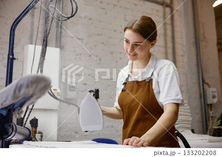 A woman worker wearing an apron is skillfully ironing a shirt on a table 133202208