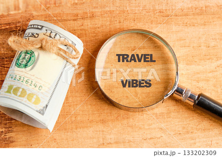 A stack of one hundred dollar bills secured with twine sits beside a magnifying glass that shows the words TRAVEL VIBES on a textured wooden surface A stack of one hundred dollar bills secured with twine sits beside a magnifying glass that shows the words TRAVEL VIBES on a textured wooden surface 133202309