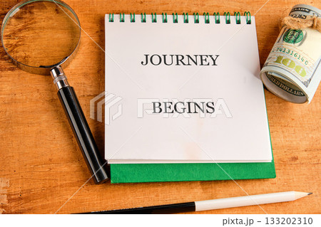 Notebook rests open on a wooden table, displaying the phrase JOURNEY BEGINS. A magnifying glass, pen, and roll of money sit nearby, suggesting planning and preparation 133202310