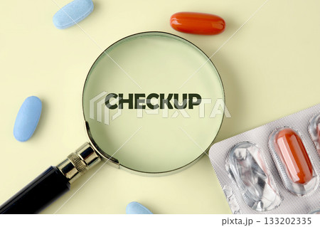 A magnifying glass focuses on the word Checkup. Around it are scattered blue pills and orange pills, some in blister packs, all on a bright neutral background 133202335