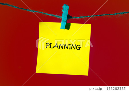 A yellow sticky note with the word Planning written in black ink is suspended from a thin blue string by a blue clothespin, set against a solid, deep red background A yellow sticky note with the word Planning written in black ink is suspended from a thin blue string by a blue clothespin, set against a solid, deep red background 133202385