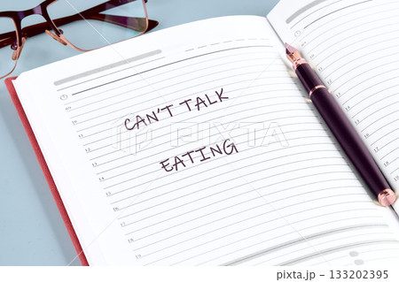 Open notebook with the words Cant Talk Eating hand-written on a lined page. A pen rests on the book with glasses in the background on the table 133202395