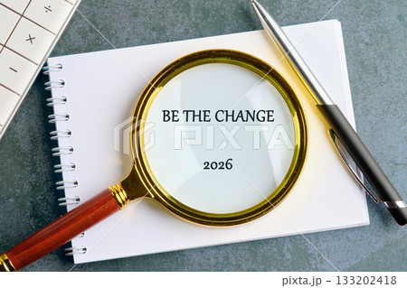 A magnifying glass with a gold rim sits on a notebook with the words BE THE CHANGE and 2026 visible. A pen rests to the side, next to a calculator 133202418