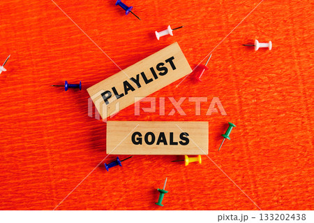 Two wooden blocks with the words playlist and goals are set against a bright orange surface, surrounded by colorful push pins, symbolizing organization and creativity in music selection 133202438