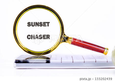 A vintage magnifying glass highlights the words Sunset Chaser on a tablet laying on top of a computer keyboard. The goal is to plan the next golden hour adventure 133202439