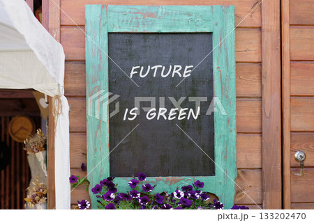 A chalkboard sign with the words FUTURE IS GREEN hangs on a wooden building. Violet flowers decorate below the sign, symbolizing a hopeful environmental message A chalkboard sign with the words FUTURE IS GREEN hangs on a wooden building. Violet flowers decorate below the sign, symbolizing a hopeful environmental message 133202470