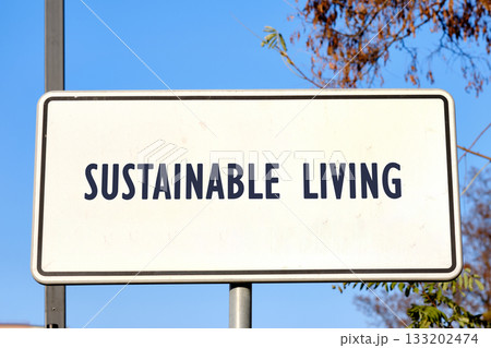 A rectangular white sign says Sustainable Living in dark blue letters, under a clear blue Autumn sky, promoting a green lifestyle near some trees A rectangular white sign says Sustainable Living in dark blue letters, under a clear blue Autumn sky, promoting a green lifestyle near some trees 133202474