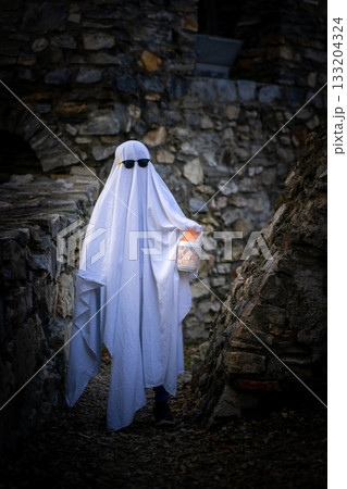 A child in a white sheet, sunglasses and a lantern in hands depicts a ghost against the background of a stone wall of an ancient building. Halloween 133204324