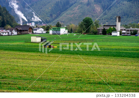 A tractor is engaged in harvesting on a small private agricultural field against the backdrop of the Alpine mountains in Austria A tractor is engaged in harvesting on a small private agricultural field against the backdrop of the Alpine mountains in Austria 133204338