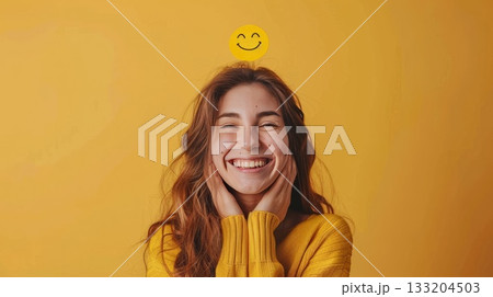 Woman in yellow smiling widely with a smiley face above her head, on a yellow background. Concept of happiness and positivity 133204503