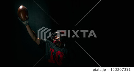 Football player raising ball upward with dramatic emotional expression on dark background. Banner 133207351