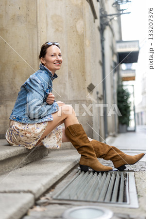 Portrait of young adult woman wear floral dress denim jacket brown suede boots  sitting on stairs outdoors in old European city. Stylish female person enjoy walk travel urban lifestyle summertime day 133207376