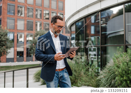 Businessman in front of office building, holding tablet and reading something. 133209151