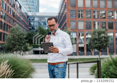 Businessman in front of office building, holding tablet and reading something. 133209152