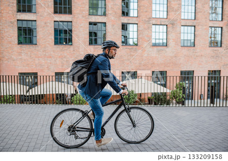 Side view of businessman riding bike to office, wearing backpack and helmet. 133209158