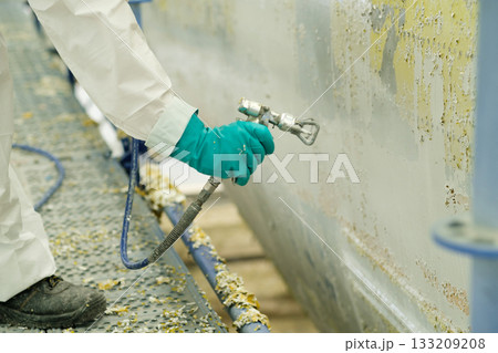 Worker in protective gear uses spray equipment to remove paint from a boat hull in a shipyard during day hours 133209208