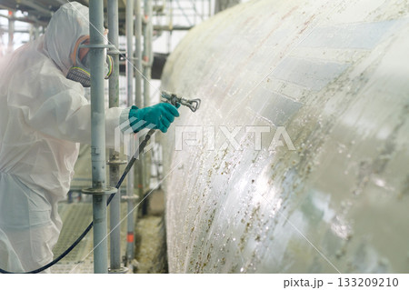 Worker in protective gear spray painting a large industrial tank at a construction site during daylight hours Worker in protective gear spray painting a large industrial tank at a construction site during daylight hours 133209210