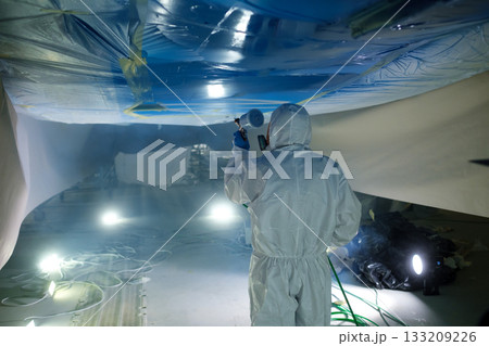 Worker in protective suit paints underside of a boat in a well-lit workshop, showcasing meticulous craftsmanship and attention to detail during a restoration project 133209226